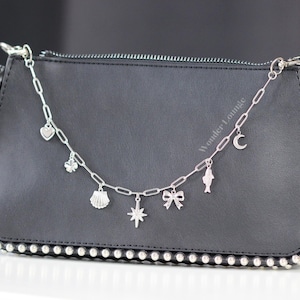 May include: A black clutch bag with silver studs around the edges. A silver chain with charms, including a heart, clover, seashell, star, bow, fish, and crescent moon, hangs from the top. The text "Wonder Lounge" is visible on the bag.