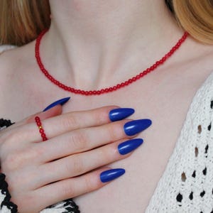 May include: A red beaded necklace with a gold clasp. The necklace is worn by a person with blue nail polish.