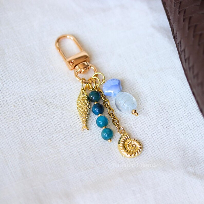 Op de afbeelding: Goudkleurige sleutelhanger met een karabijnhaak, met een visbedel, blauwe kralen en een spiraalvormige schelpenhanger. De sleutelhanger bevat ook een ketting en lichtblauwe en witte edelsteenaccenten. De achtergrond is een witte stof.