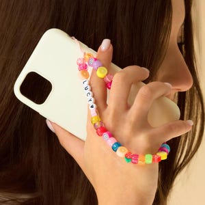 May include: A white iPhone case with a colorful beaded phone strap. The strap has a heart and the name "LUCY" spelled out in white beads.