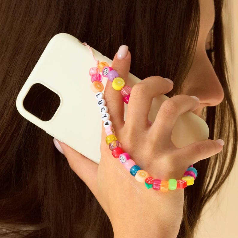 May include: A white iPhone case with a colorful beaded phone strap. The strap has a heart and the name "LUCY" spelled out in white beads.