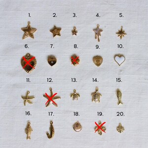 May include: A collection of gold-colored charms in various shapes, including stars, hearts, a turtle, a fish, and a seashell. Some charms have a textured surface, while others are smooth. The charms are arranged on a light-colored surface.