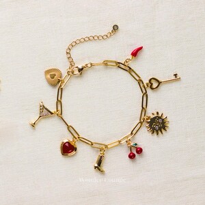 May include: Gold-toned charm bracelet featuring various charms, including a martini glass, heart, boot, cherries, key, sun, and a red chili pepper. The bracelet is displayed on a white surface.