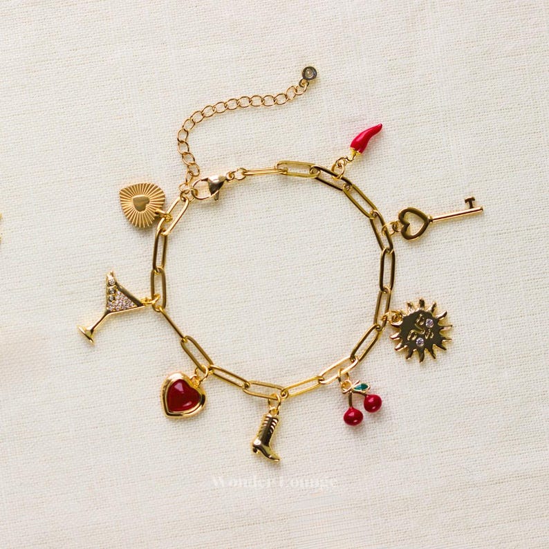 May include: Gold-toned charm bracelet featuring various charms, including a martini glass, heart, boot, cherries, key, sun, and a red chili pepper. The bracelet is displayed on a white surface.