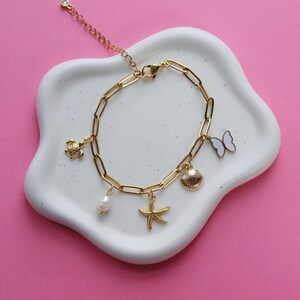 Peut inclure: Un bracelet en cha&icirc;ne dor&eacute;e avec des breloques en forme de tortue, de papillon, de coquillage, d'&eacute;toile de mer et de perle. Le bracelet est sur un plat en c&eacute;ramique blanche.