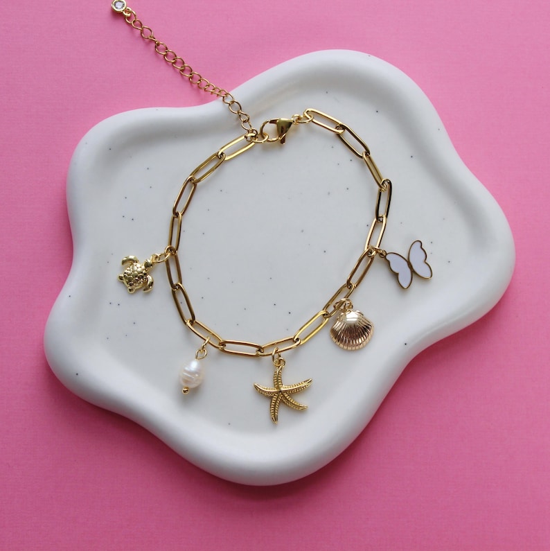 Peut inclure: Un bracelet en cha&icirc;ne dor&eacute;e avec des breloques en forme de tortue, de papillon, de coquillage, d'&eacute;toile de mer et de perle. Le bracelet est sur un plat en c&eacute;ramique blanche.