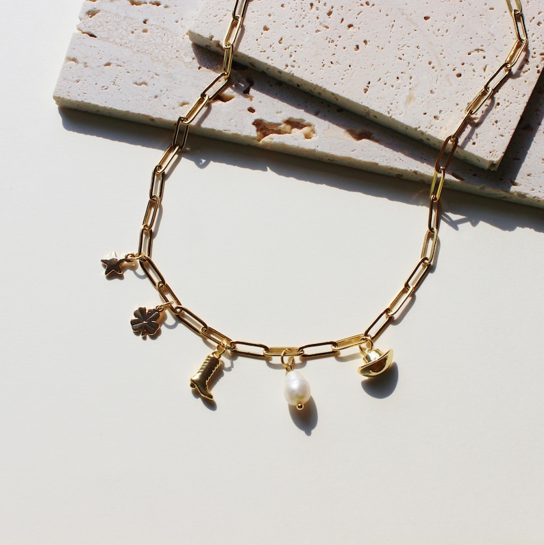 Puede incluir: Un collar de cadena dorada con varios colgantes, incluyendo una estrella, un tr&eacute;bol de cuatro hojas, una bota, una perla y un sombrero de vaquero.