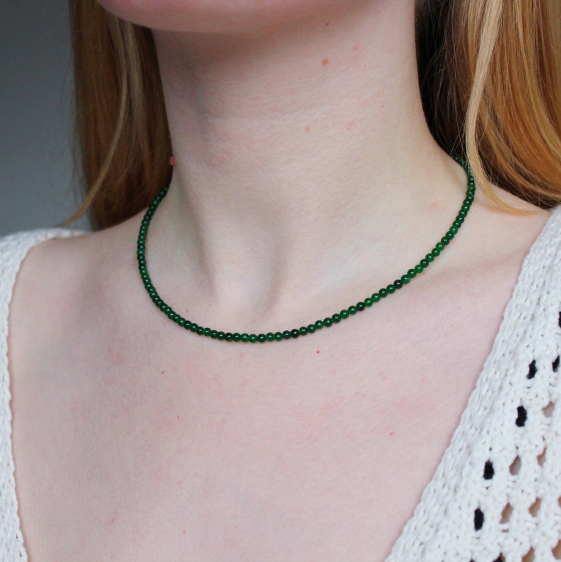 May include: A delicate green jade necklace with small round beads. The necklace is worn by a person with a white crocheted top.