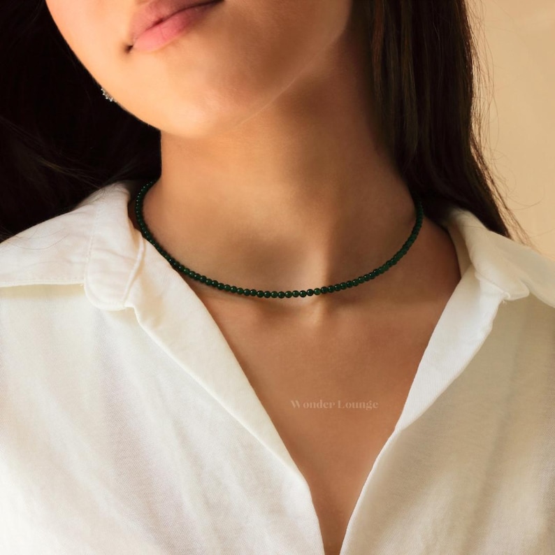 May include: A delicate choker necklace made of small, dark green beads. The necklace is worn by a person with a white shirt.