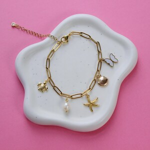 Peut inclure: Un bracelet en cha&icirc;ne dor&eacute;e avec plusieurs breloques, dont une tortue, un coquillage, une &eacute;toile de mer, une perle et un papillon. Le bracelet est sur un fond blanc.