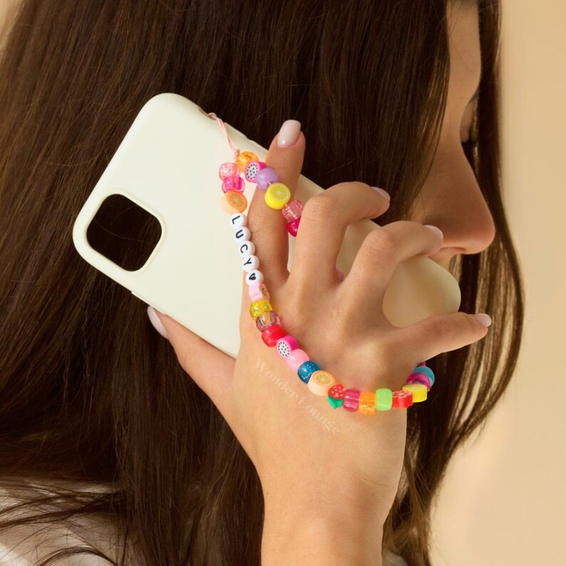 May include: A white phone case with a colorful beaded phone strap. The strap has the name "LUCY" spelled out in white beads.