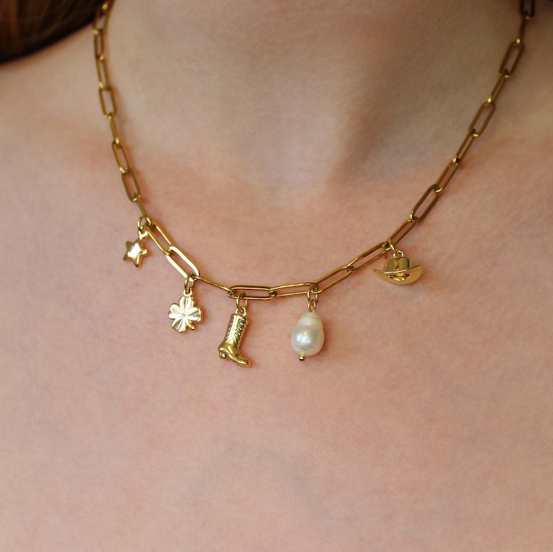 Puede incluir: Un collar de cadena dorada con m&uacute;ltiples dijes, incluyendo una estrella, un tr&eacute;bol de cuatro hojas, una bota, una perla y un sombrero de vaquero.