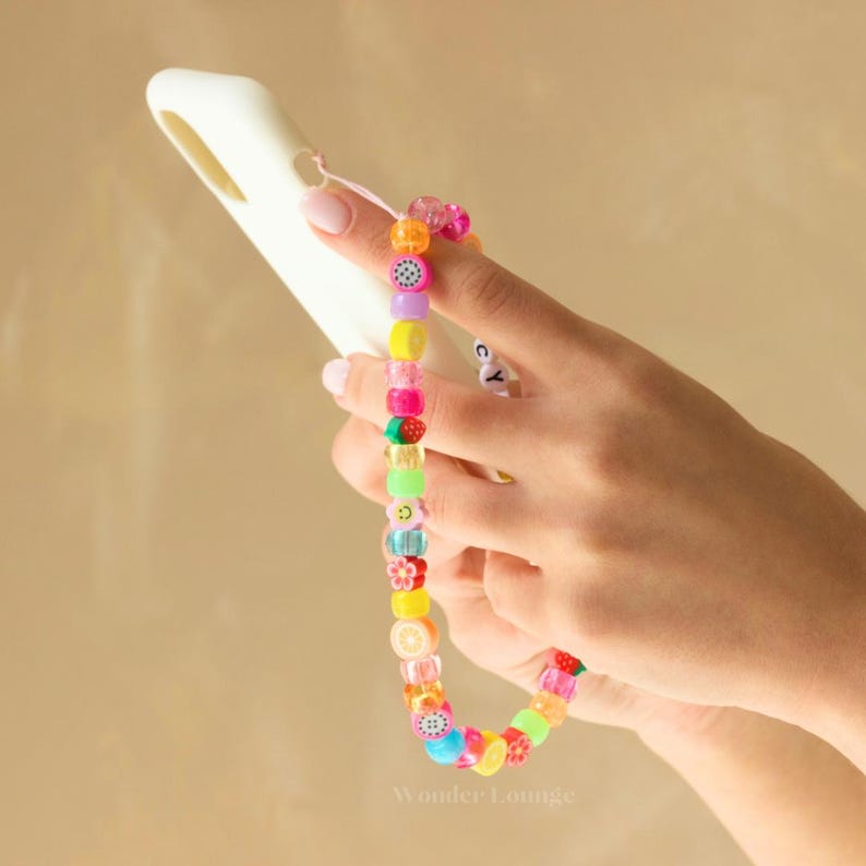 May include: A colorful beaded phone strap with fruit and smiley face charms. The strap is attached to a white phone case.