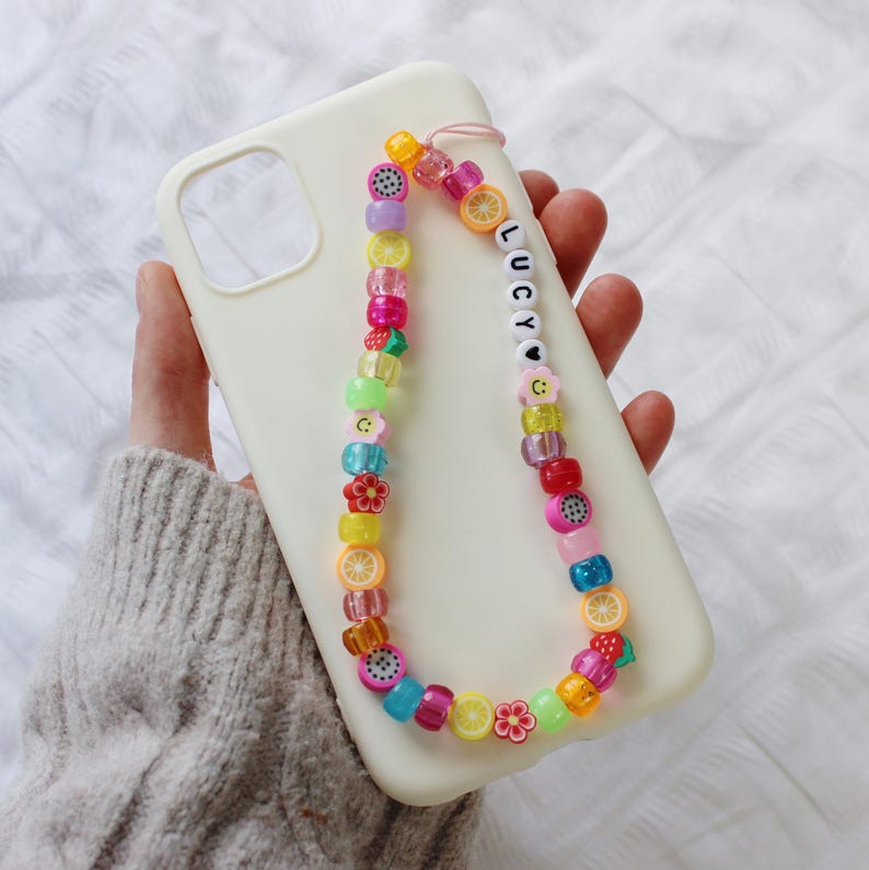 May include: A white phone case with a colorful beaded phone strap. The strap features fruit and flower charms and the name "LUCY" spelled out in white beads.