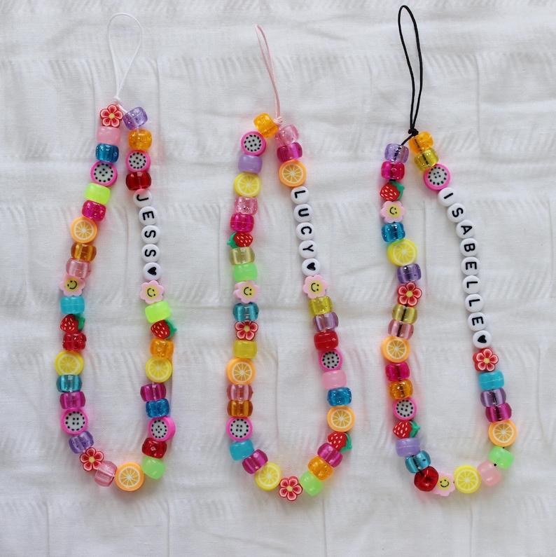 May include: Three colorful beaded phone charms with the names "Jess", "Lucy", and "Isabelle" spelled out in white beads. Each charm features a variety of fruit and flower beads in pink, yellow, green, blue, and purple.