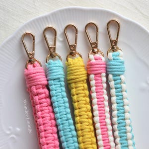 May include: Five macrame keyrings in pink, blue, yellow, pink and white striped, and blue and white striped patterns. Each keyring has a gold-coloured clasp. The keyrings are arranged on a white plate.