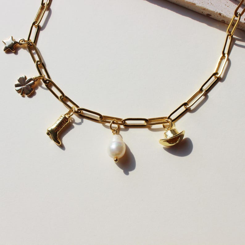 Puede incluir: Un collar de cadena dorada con m&uacute;ltiples dijes, incluyendo una estrella, un tr&eacute;bol de cuatro hojas, una bota, una perla y un sombrero de vaquero.