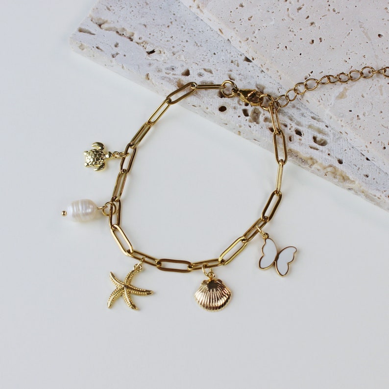Peut inclure: Un bracelet en cha&icirc;ne dor&eacute;e avec des breloques, dont un coquillage, une &eacute;toile de mer, un papillon, une tortue et une perle.