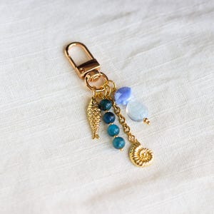 Op de afbeelding: Goudkleurige sleutelhanger met een karabijnhaak, met een visbedel, blauwe kralen en een schelpenbedel. De sleutelhanger heeft een gouden ketting en is versierd met lichtblauwe en transparante kralen.