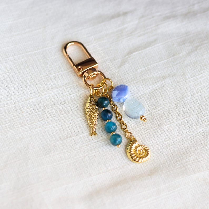 Op de afbeelding: Goudkleurige sleutelhanger met een karabijnhaak, met een visbedel, blauwe kralen en een schelpenbedel. De sleutelhanger heeft een gouden ketting en is versierd met lichtblauwe en transparante kralen.