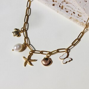 Peut inclure: Un collier en cha&icirc;ne dor&eacute;e avec des breloques, dont une perle, un coquillage, une &eacute;toile de mer, un papillon et une tortue. Les breloques sont toutes dor&eacute;es et ont une finition &eacute;maill&eacute;e blanche.