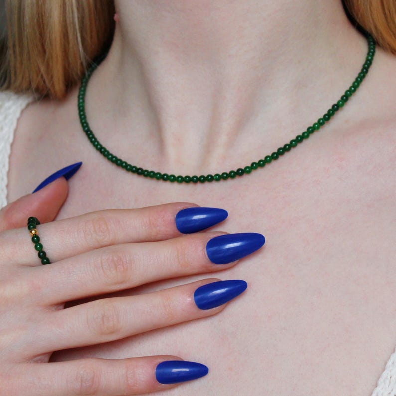 May include: A green jade beaded necklace with a delicate design. The necklace is worn by a person with blue nail polish.