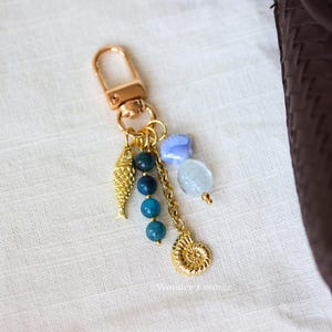 Op de afbeelding: Goudkleurige sleutelhanger met een karabijnhaak, met een visbedel, blauwe kralen en een schelpenbedel. Extra bedels zijn een blauwe schelp en een heldere, ovale kraal. De sleutelhanger heeft een nautisch thema.