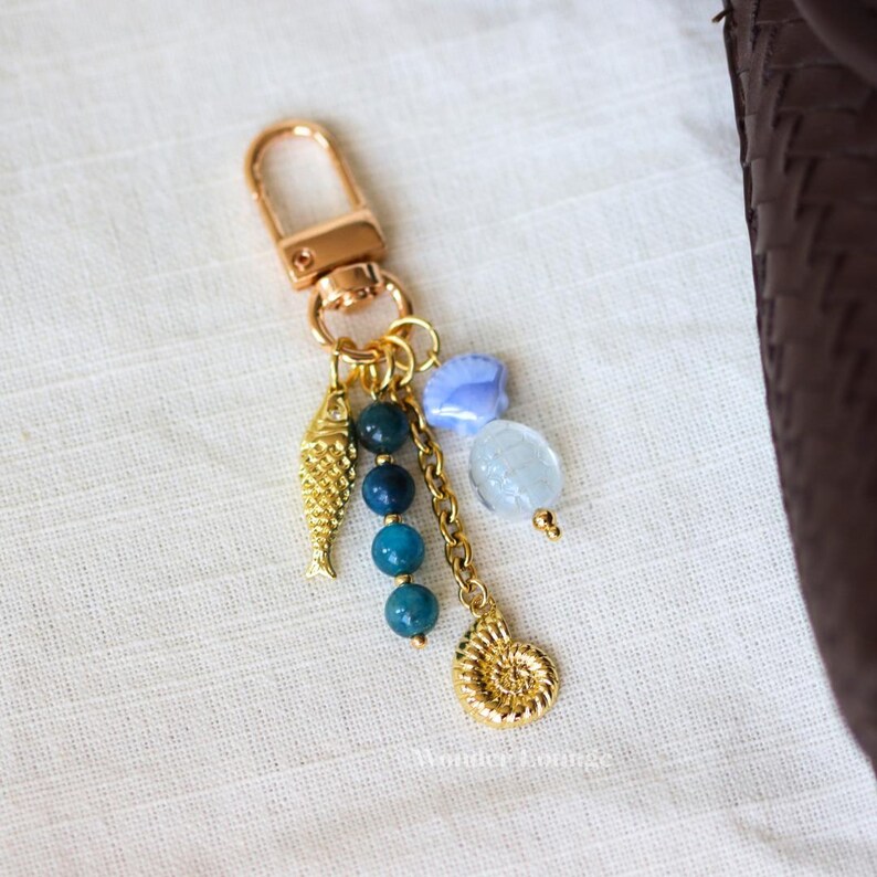 Op de afbeelding: Goudkleurige sleutelhanger met een karabijnhaak, met een visbedel, blauwe kralen en een schelpenbedel. Extra bedels zijn een blauwe schelp en een heldere, ovale kraal. De sleutelhanger heeft een nautisch thema.