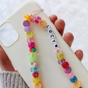 May include: A white phone case with a colorful beaded phone strap. The strap features fruit charms, smiley faces, and the name "Lucy" spelled out in white beads.