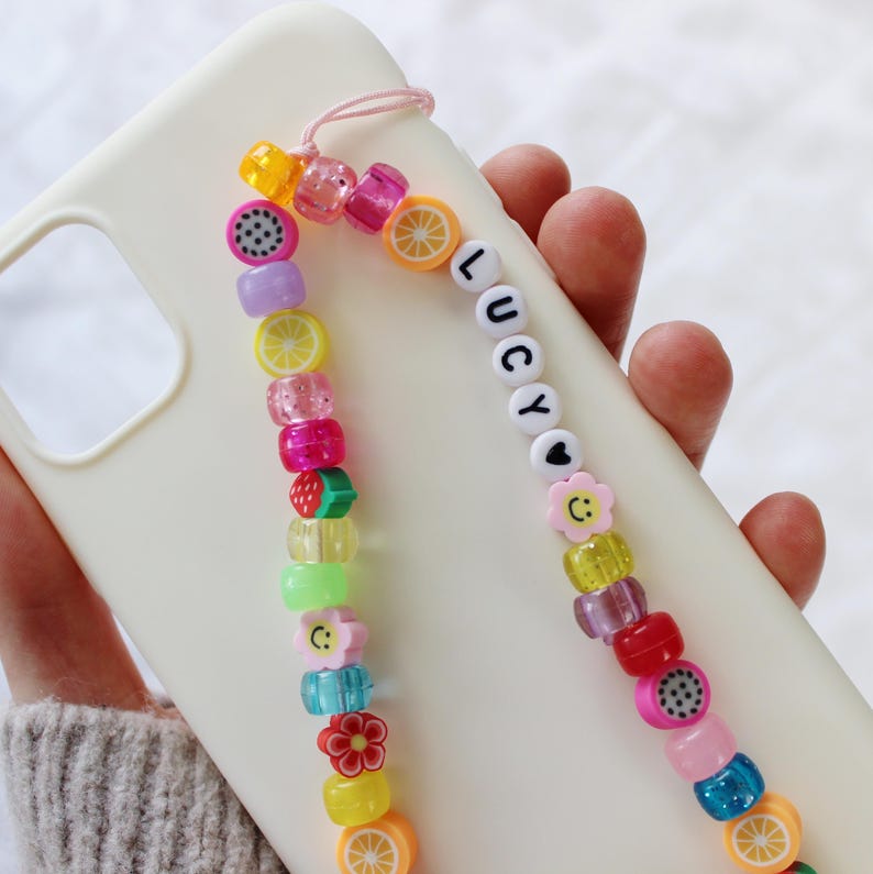 May include: A white phone case with a colorful beaded phone strap. The strap features fruit charms, smiley faces, and the name "Lucy" spelled out in white beads.