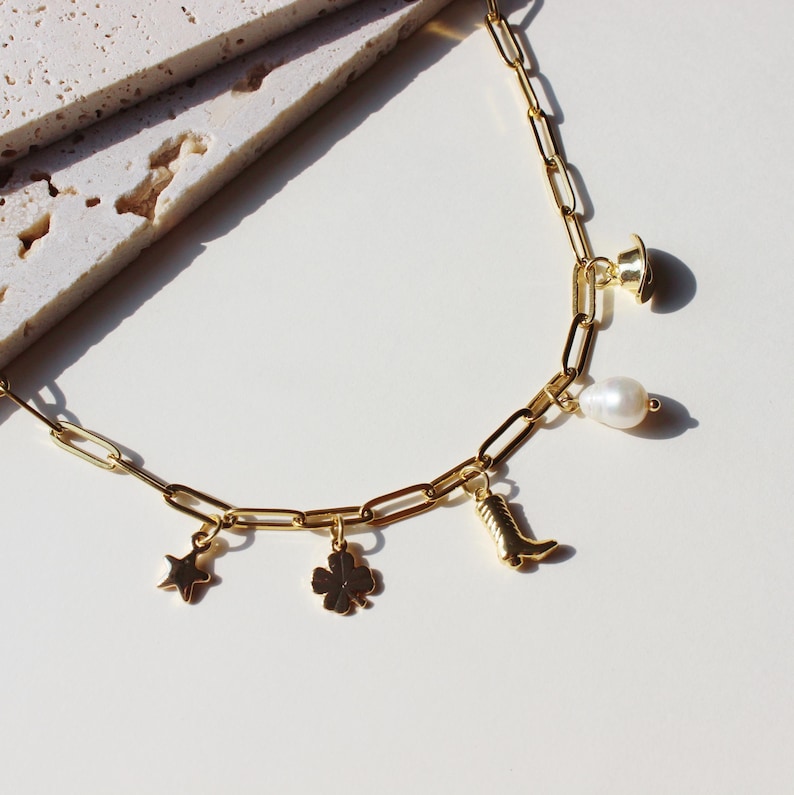 Puede incluir: Un collar de cadena dorada con m&uacute;ltiples dijes, incluyendo una estrella, un tr&eacute;bol de cuatro hojas, una bota, una perla y un peque&ntilde;o sombrero.