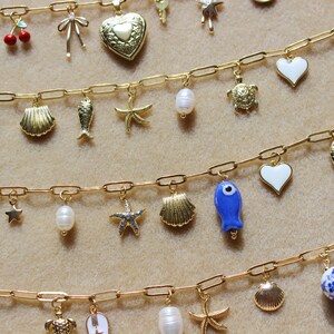 May include: Gold-toned charm bracelets with various pendants, including seashells, starfish, pearls, and a blue fish. The charms are attached to a delicate chain. The background is a neutral beige.