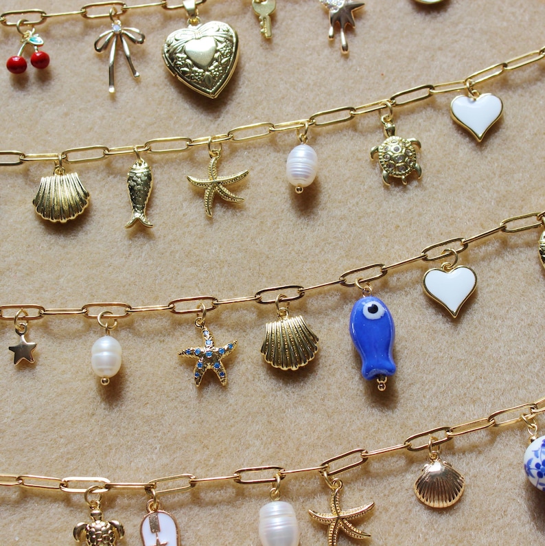 May include: Gold-toned charm bracelets with various pendants, including seashells, starfish, pearls, and a blue fish. The charms are attached to a delicate chain. The background is a neutral beige.