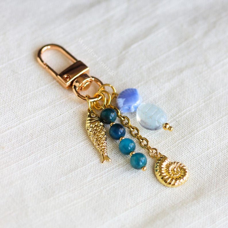 Op de afbeelding: Goudkleurige sleutelhanger met een karabijnhaak, met een kleine, getextureerde vissenbedel, blauwe kralen en een spiraalvormige schelpenbedel. Extra bedels zijn een blauwe zeeschelp en een lichtblauwe ovale kraal. De sleutelhanger heeft een nautisch thema.