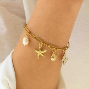 Peut inclure: Bracelet en cha&icirc;ne dor&eacute;e avec des breloques, dont une &eacute;toile de mer, une coquille, une perle et un papillon. Le bracelet est port&eacute; au poignet.