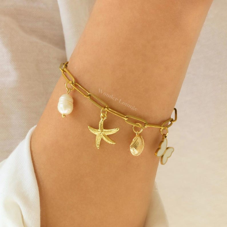 Peut inclure: Bracelet en cha&icirc;ne dor&eacute;e avec des breloques, dont une &eacute;toile de mer, une coquille, une perle et un papillon. Le bracelet est port&eacute; au poignet.