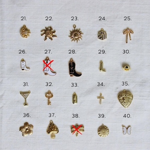 May include: A collection of gold-colored charms, including a sunflower, spider, sun, bow, cowboy boots, martini glass, key, cross, heart locket, flower, mushroom, and butterfly. Each charm is numbered from 21 to 40.