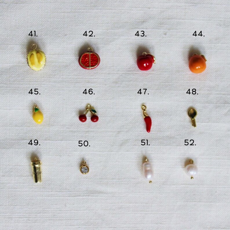 May include: A collection of small, colorful charms in various shapes, including fruits like a durian, pomegranate, apple, and orange. Other charms include a lemon, cherries, a chili pepper, a key, a small cup, a pendant with a gem, and two pearl charms.