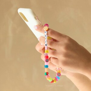 May include: A colorful beaded phone strap with the name "Lucy" spelled out in white beads. The strap is attached to a white phone case.