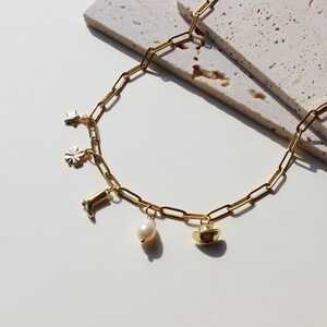 Puede incluir: Un collar de cadena dorada con m&uacute;ltiples dijes, incluyendo una estrella, una flor, una bota, una perla y un sombrero de vaquero.