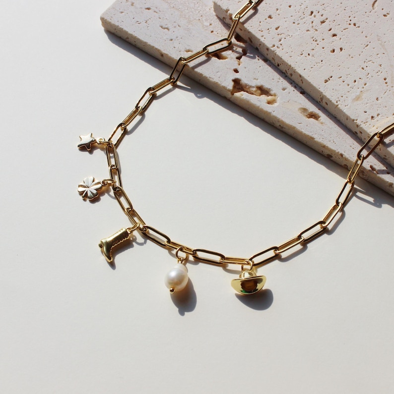 Puede incluir: Un collar de cadena dorada con m&uacute;ltiples dijes, incluyendo una estrella, una flor, una bota, una perla y un sombrero de vaquero.