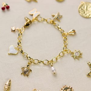 May include: Gold-toned charm bracelet featuring various pendants, including a white heart, a butterfly, a star, a sunburst, a turtle, and a pearl. The bracelet has an oval link chain and a lobster clasp.