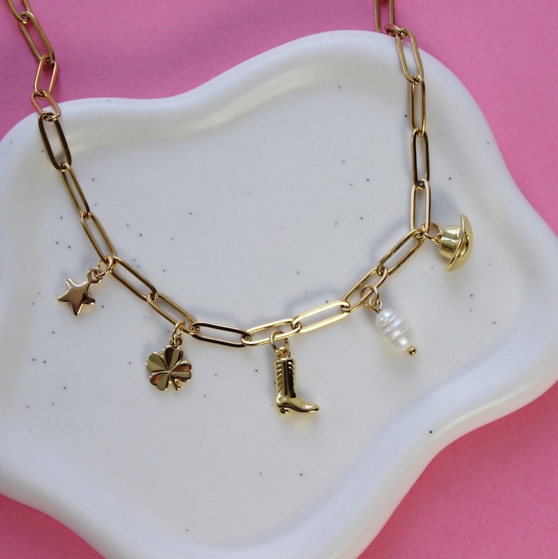 Puede incluir: Un collar de cadena dorada con varios colgantes, incluyendo una estrella, un tr&eacute;bol de cuatro hojas, una bota, una perla y un sombrero de vaquero.