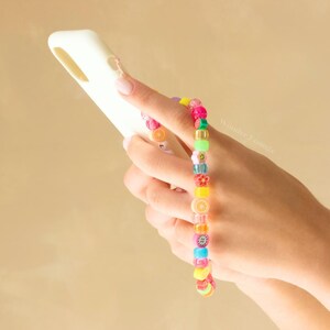 May include: A colorful beaded bracelet with fruit and flower charms. The bracelet is worn on the wrist of a person holding a white phone case.