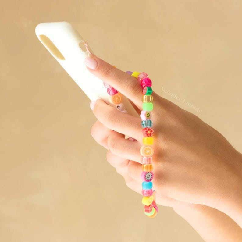 May include: A colorful beaded bracelet with fruit and flower charms. The bracelet is worn on the wrist of a person holding a white phone case.