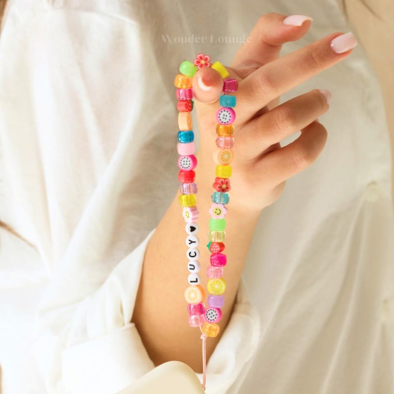 May include: A colorful phone charm with a beaded chain featuring fruit and flower designs. The charm also includes the name "LUCY" spelled out in white beads with a black heart.