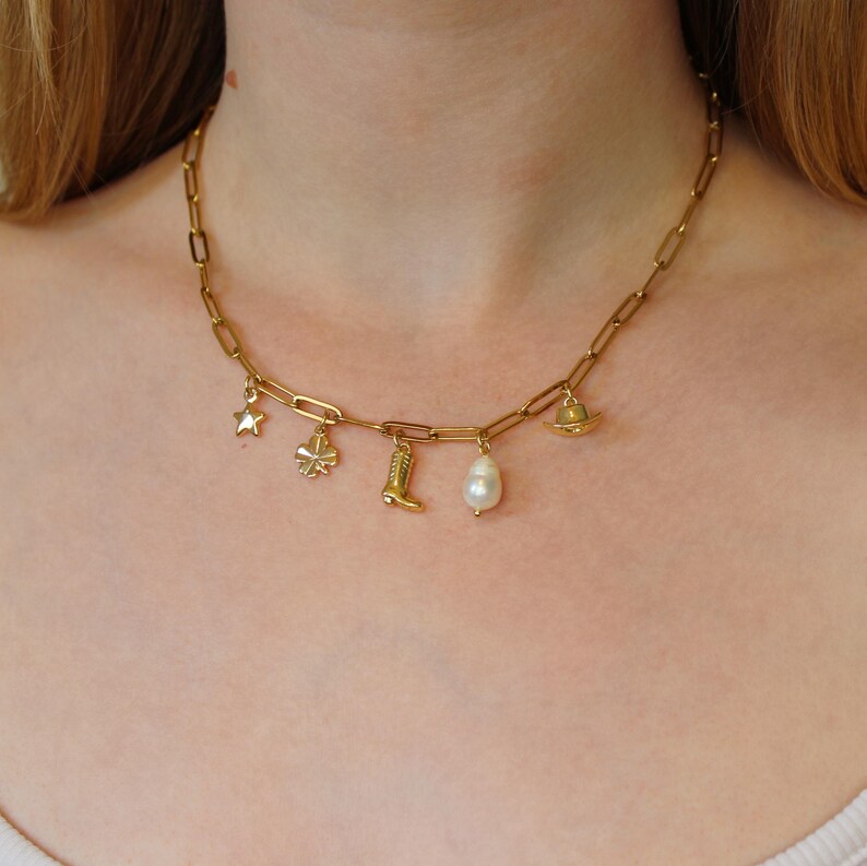 Puede incluir: Un collar de cadena dorada con varios colgantes, incluyendo una estrella, un tr&eacute;bol de cuatro hojas, una bota, una perla y un peque&ntilde;o sombrero.
