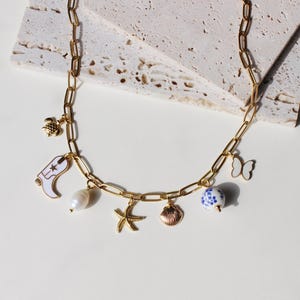 May include: Gold-tone chain necklace with various charms, including a white cowboy boot, a pearl, a starfish, a seashell, a blue and white bead, a butterfly, and a turtle. The necklace is displayed on a white surface.