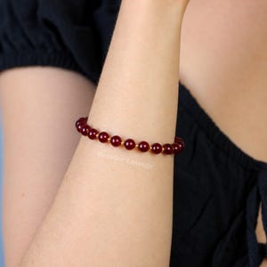 May include: A beaded bracelet featuring alternating dark red spherical beads and small gold-toned metallic spacers. The bracelet is worn on an arm, and the text Wonder Lounge is visible in white lettering near the jewelry piece.