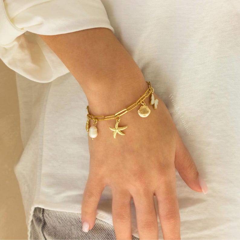 Peut inclure: Bracelet en or avec des breloques, dont une &eacute;toile de mer, des coquillages et une perle. Le bracelet est port&eacute; au poignet.